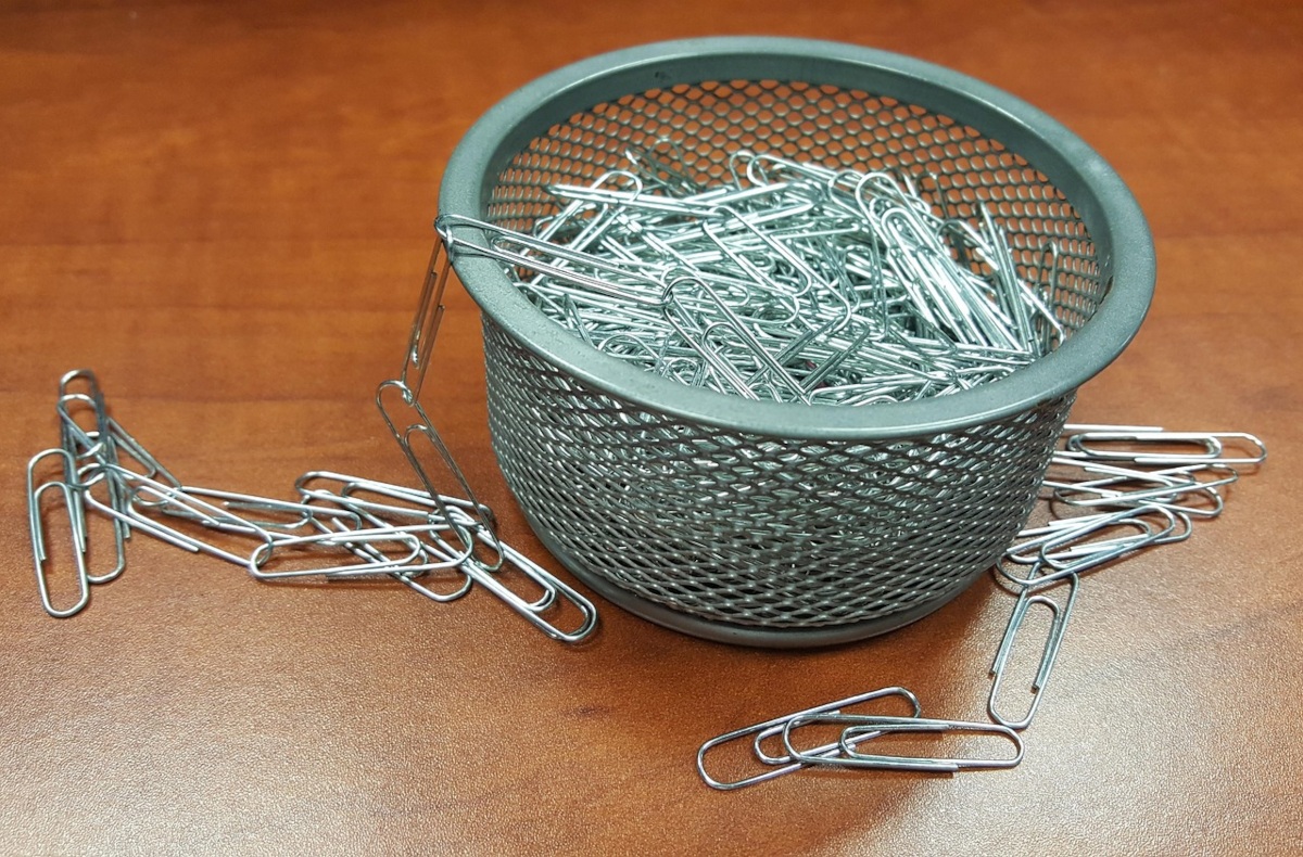 A grey mesh wire container filled to the brim with silver paperclips sitting on a wooden table, with loose paperclips spilling out onto the surface.